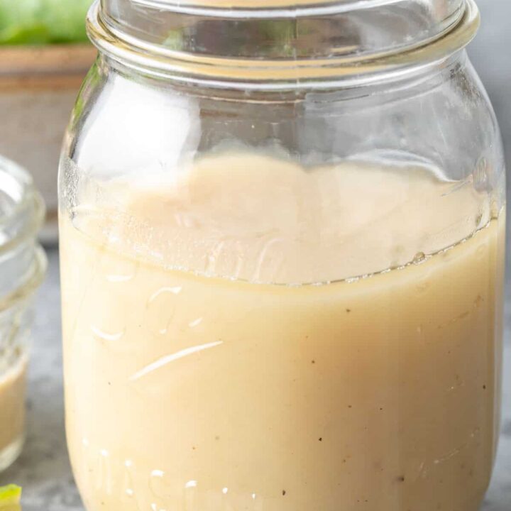 A clear glass mason jar filled with Honey Lime Tahini Dressing. A tan colored bowl filled with salad sits next to the jar.