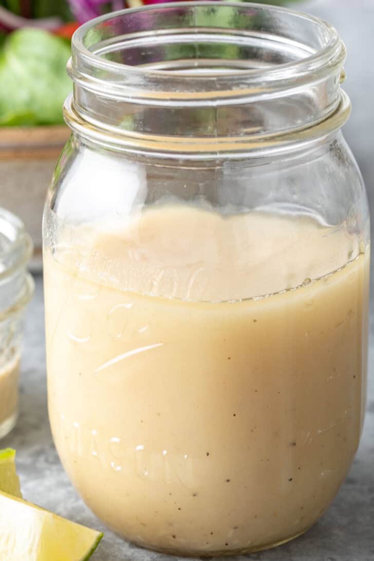 A clear glass mason jar filled with Honey Lime Tahini Dressing. A tan bowl filled with salad sits next to the jar.