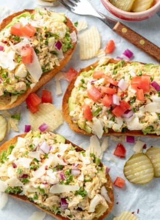 Three pieces of Tuna Avocado Toast on deli paper with chips and pickle slices scattered next to them.