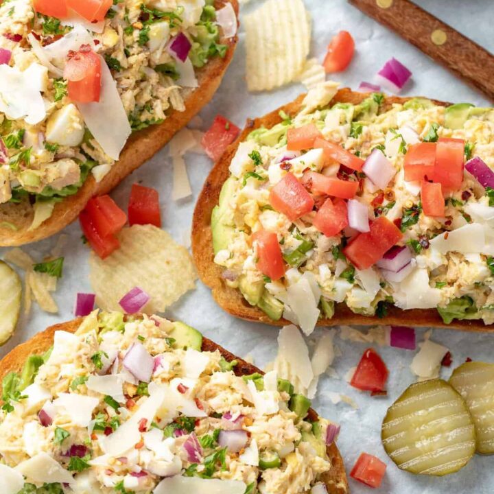 Three pieces of Tuna Avocado Toast on deli paper with chips and pickle slices scattered next to them.