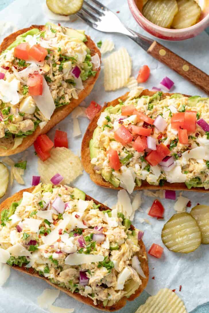 Three pieces of tuna avocado toast on deli wrap paper. Potato chips and pickles are scattered next to the toast.