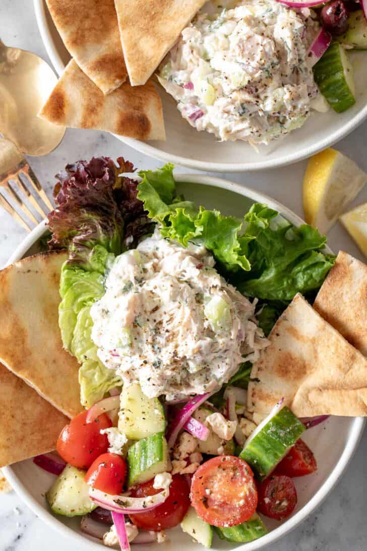 Two white plates filled with Tzatziki Chicken Salad, pieces of pita bread and a cucumber tomato salad.