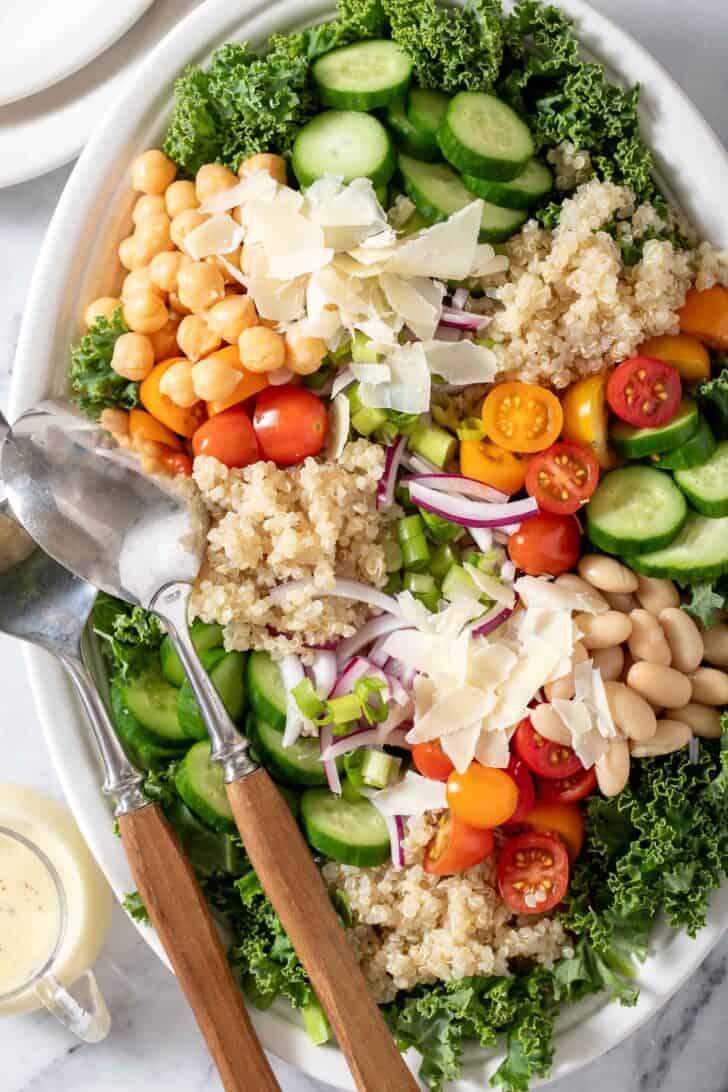 A white platter filled with curly kale, tomatoes, chickpeas and cucumber. Serving spoons with wooden handles rest on the platter.