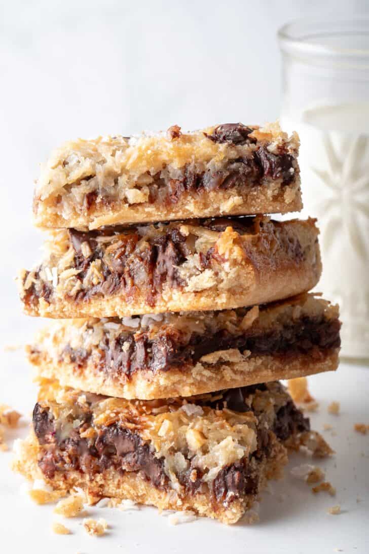 A stack of four square pieces of Magic Cookie Bars. A small glass of milk sits next to the stack.