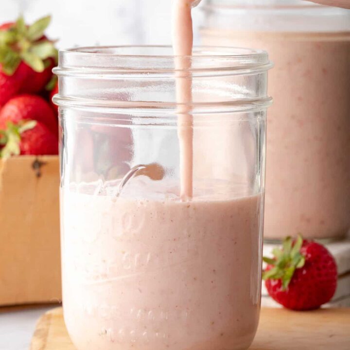 A clear glass mason jar half filled with Strawberry Banana Smoothie. Smoothie is being poured into the mason jar from a glass pitcher.