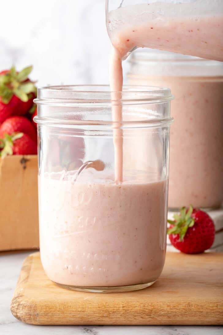 A clear glass mason jar half filled with Strawberry Banana Smoothie. Smoothie is being poured into the mason jar from a glass pitcher.