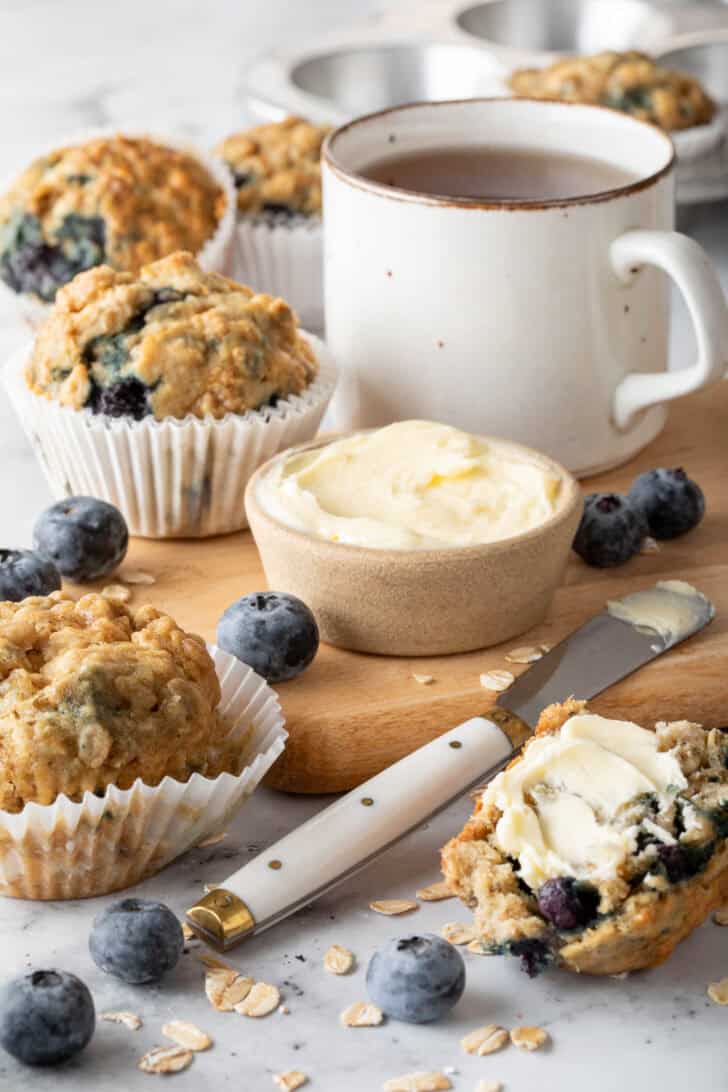 Several blueberry oatmeal muffins on a cutting board with a ramekin filled with butter.