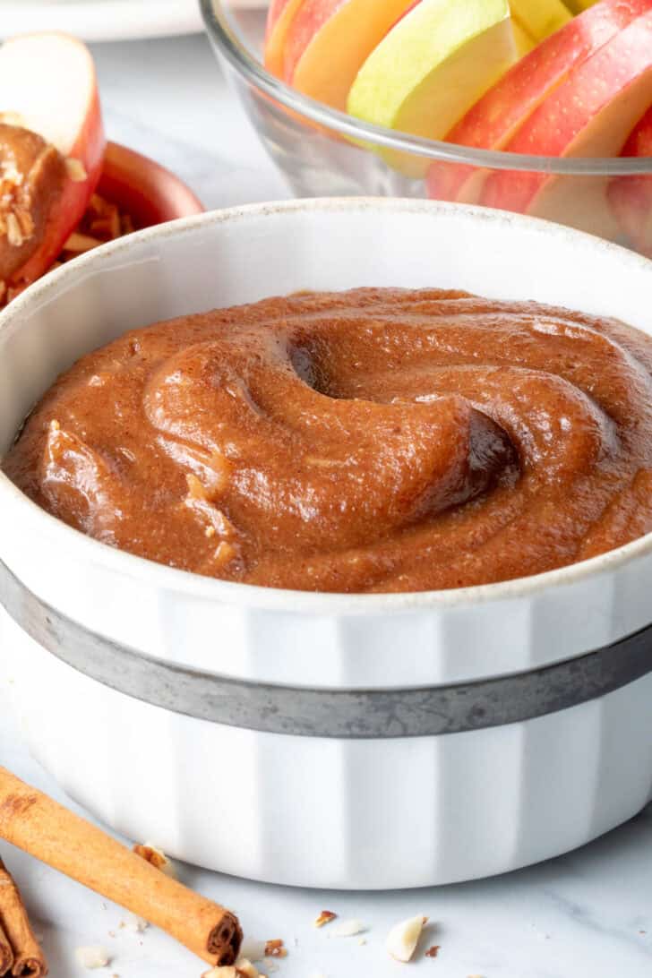 A white bowl filled with Medjool Date Caramel Sauce. A clear glass bowl filled with apple slices sits next to the bowl.