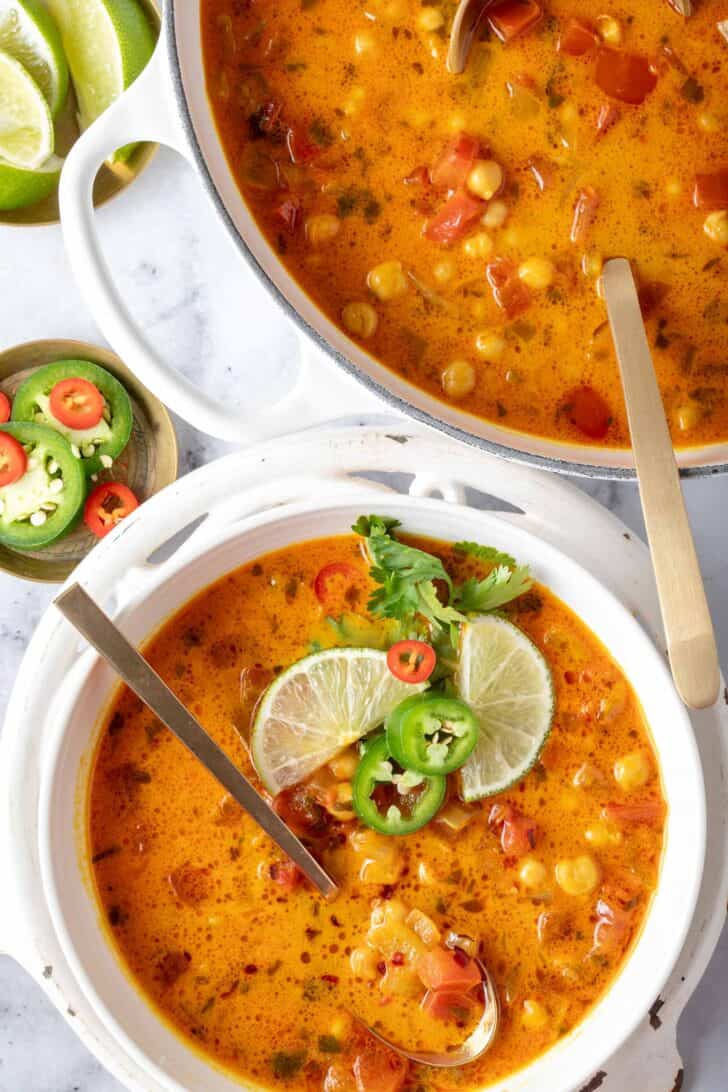 A white bowl filled with coconut chickpea curry soup sits next to a big white pot filled with the same soup.