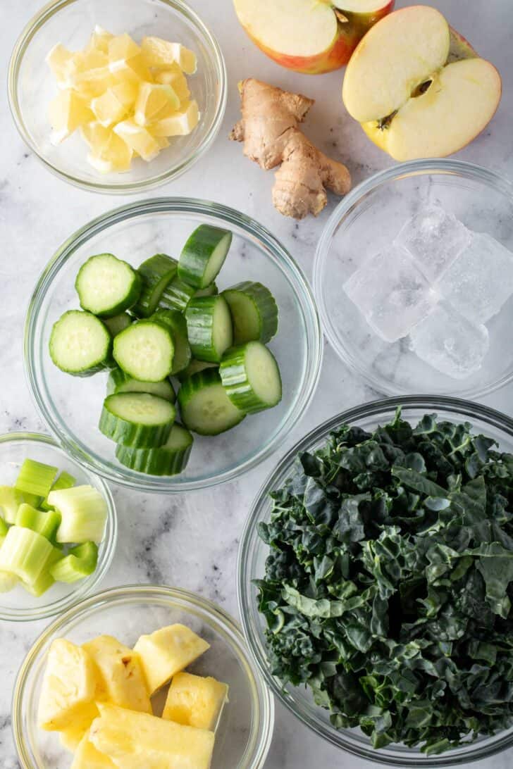 Clear glass bowls filled with chopped kale, cucumber, celery, pineapple lemon and apple.