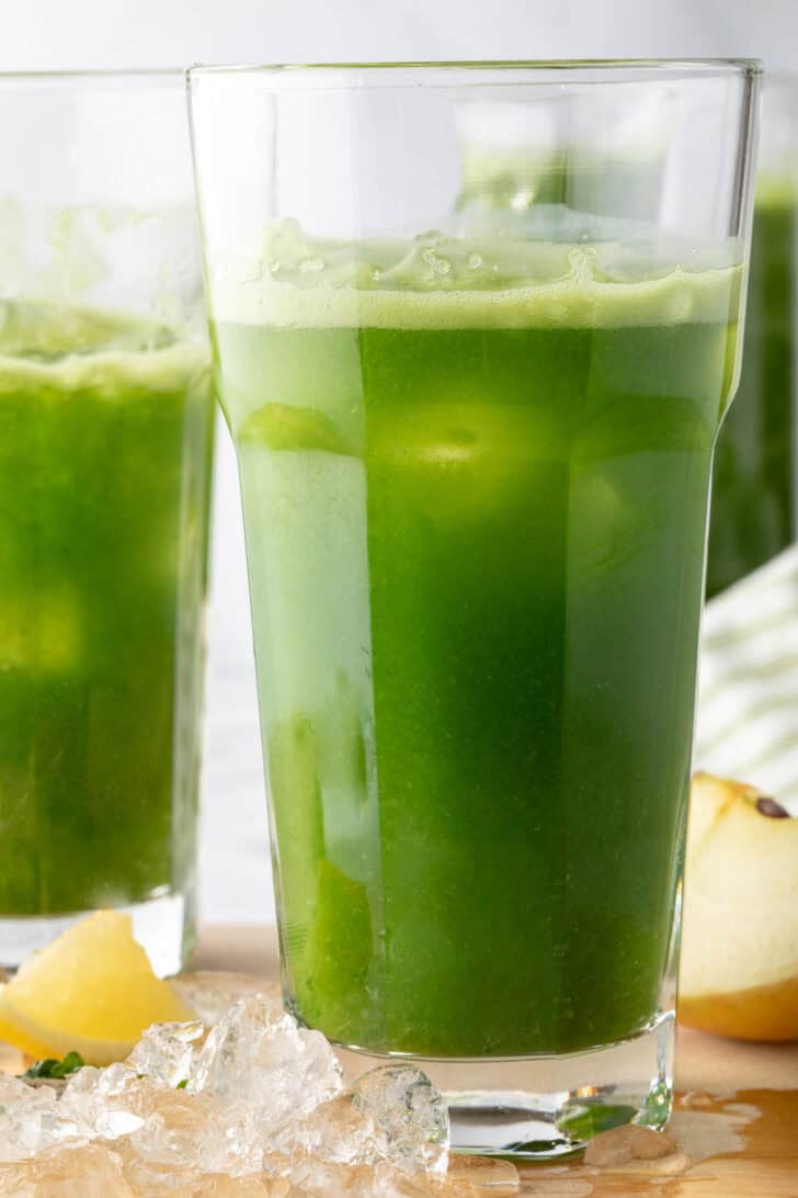 A tall clear glass filled with green juice over ice.