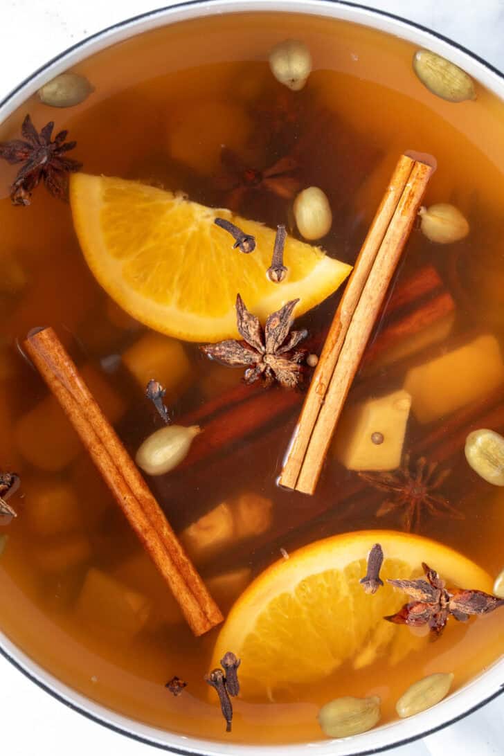 A white pot filled with cinnamon sticks, star anise, fresh ginger and orange slices simmering in water to make a tea.