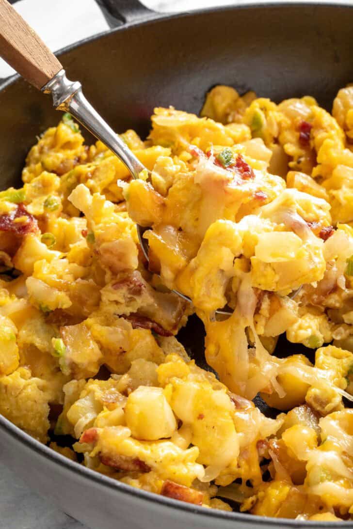 A black skillet filled with Breakfast Scramble with Potatoes. A spoon rests in the skillet.