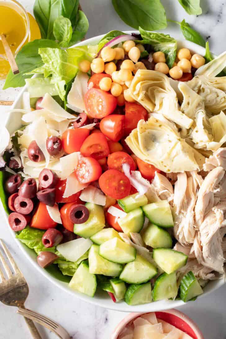 A white bowl filled with chopped lettuce, cucumber, tomato, artichoke hearts and olives.