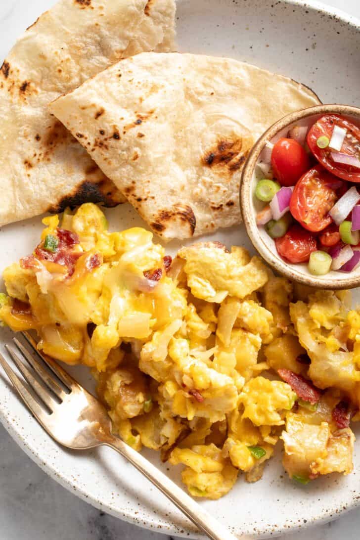 A white plate filled with breakfast scramble with potatoes. Flour tortillas sit next to the scramble.