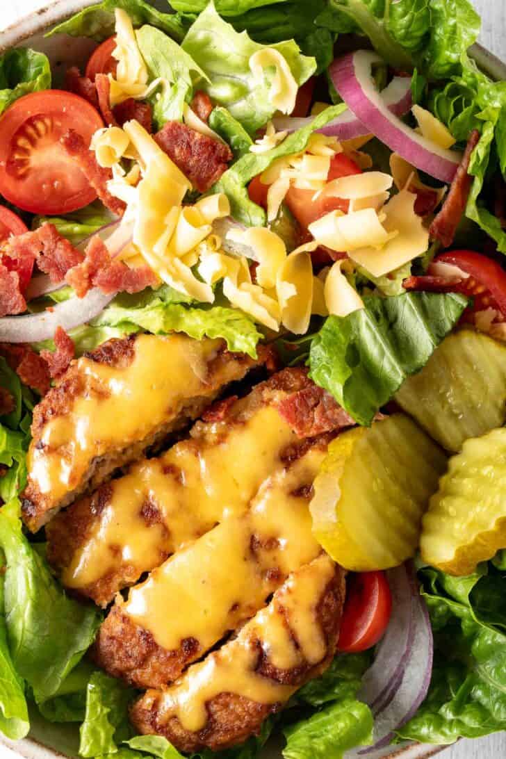 A white bowl filled with chopped lettuce, tomatoes, red onion, pickles and a ground meat patty with melted cheese.