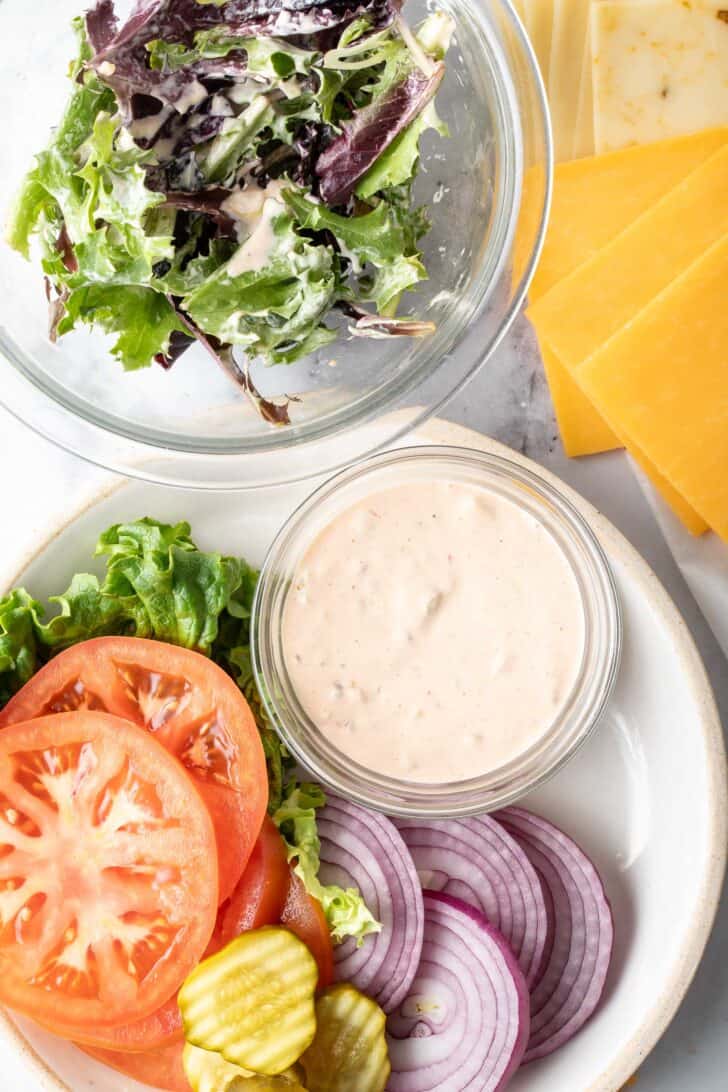 A plate with sliced tomatoes, onion and pickles next to a bowl of mixed greens tossed in dressing.