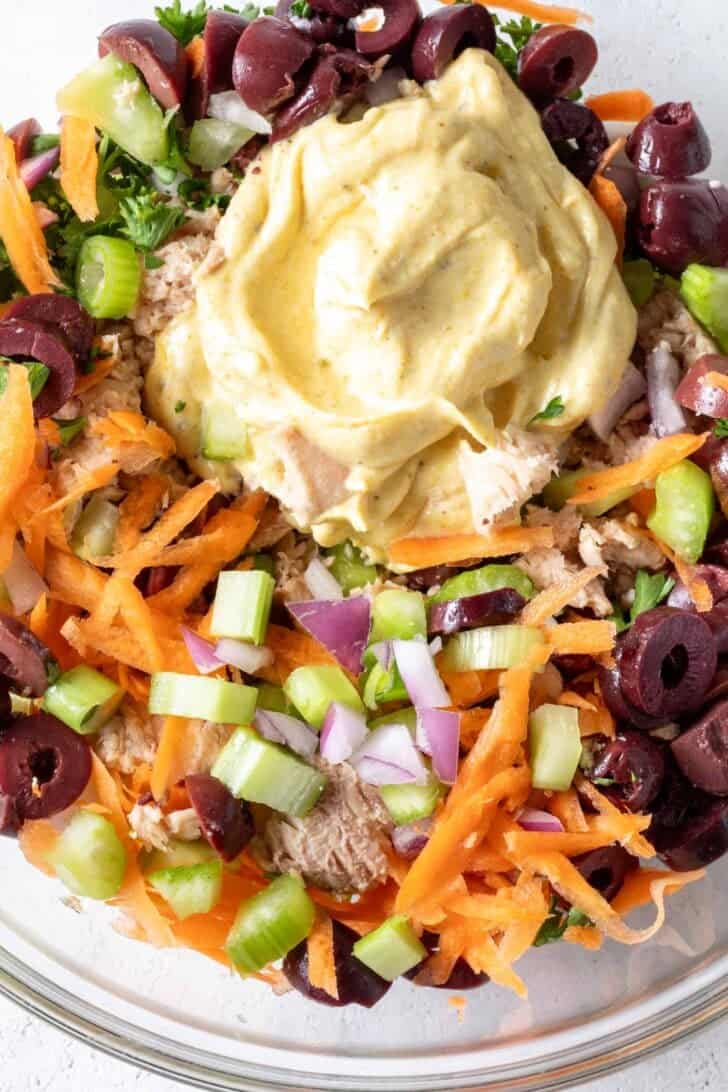 A clear glass mixing bowl filled with canned fish, chopped vegetables and a creamy dressing.