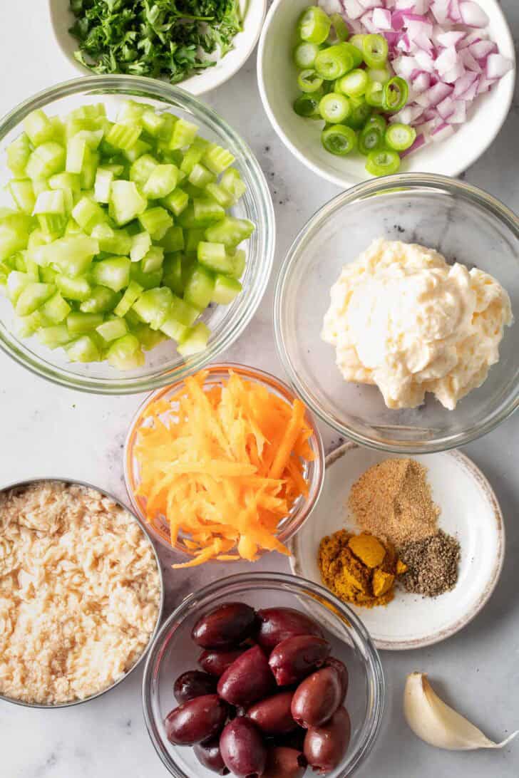 Clear glass bowls filled with canned fish, grated carrot, chopped celery, chopped onion, kalamata olives and spices.