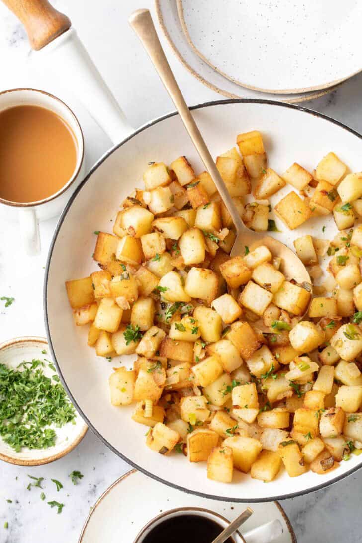 A white skillet filled with crispy breakfast potatoes. A spoon rests in the skillet.