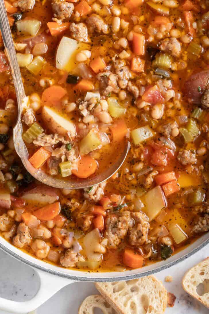 A white pot filled with Sausage Kale and White Bean Soup. A ladle rests in the pot.